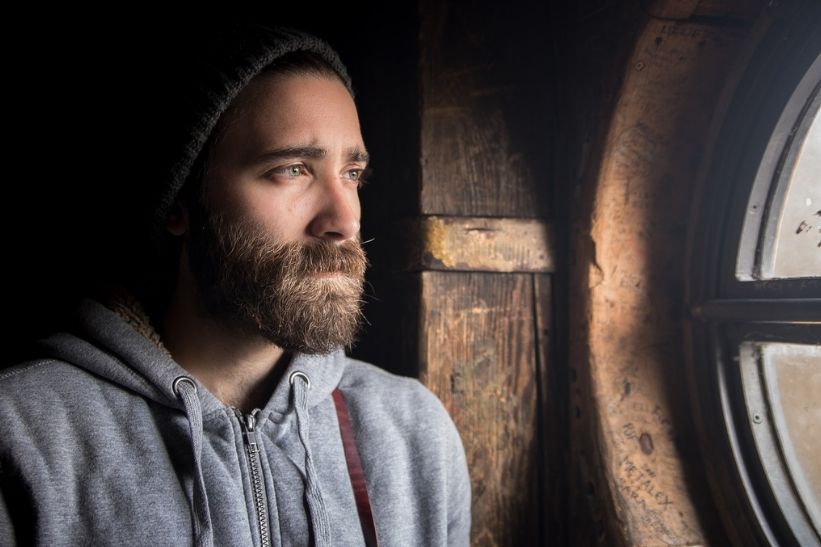 Cómo superar el miedo a vivir solo. Mi Camino a ser independiente. Foto de hombre joven con barba, mirando con preocupación a través de una pequeña ventana redonda.